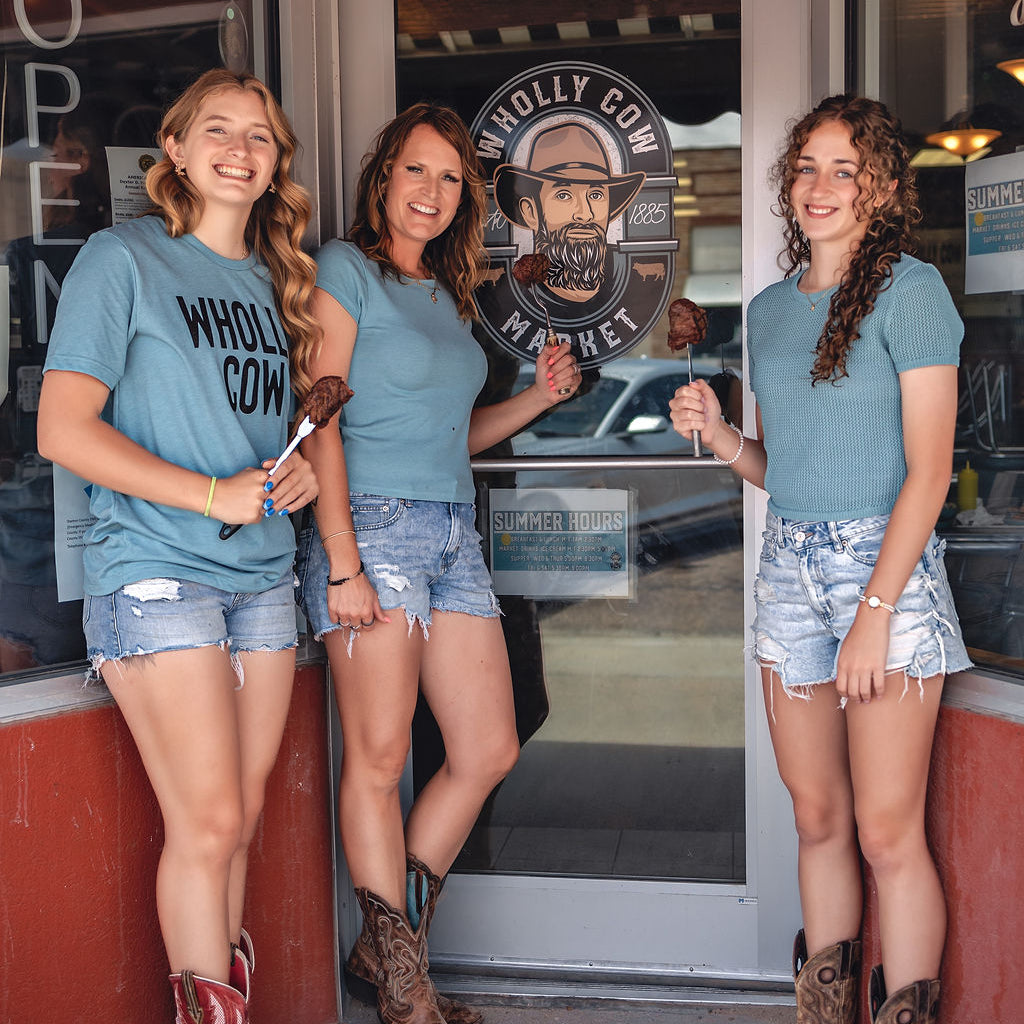 Three women in matching blue t-shirts and denim shorts standing outside a building with a 'Wholly Cow Market' sign.