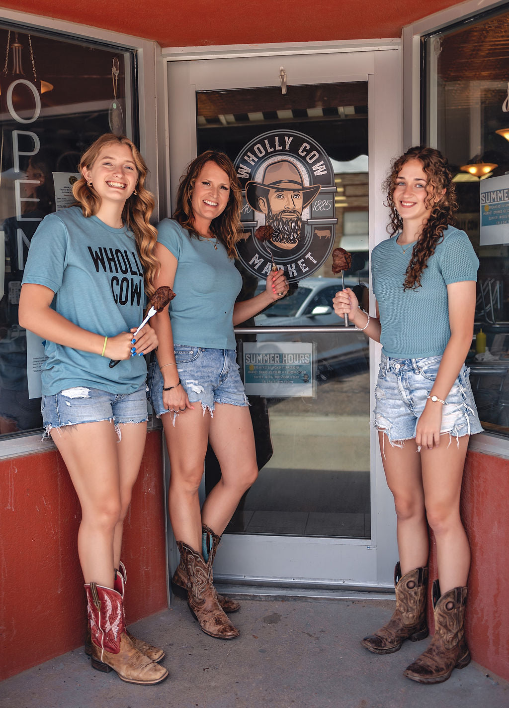 Three women in matching blue t-shirts and denim shorts standing outside a building with a 'Wholly Cow Market' sign.